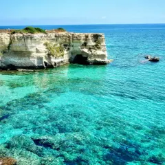 Turquoise water laps against white cliffs with caves, a boat sails in the distance.