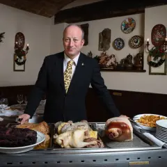 A man in a suit stands behind a cart with various meats, including ribs, a whole chicken, and a pig's head.