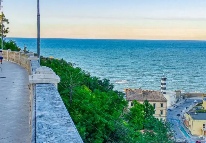 Coastal town with a lighthouse and buildings overlooking the blue sea, with a walkway in the foreground.