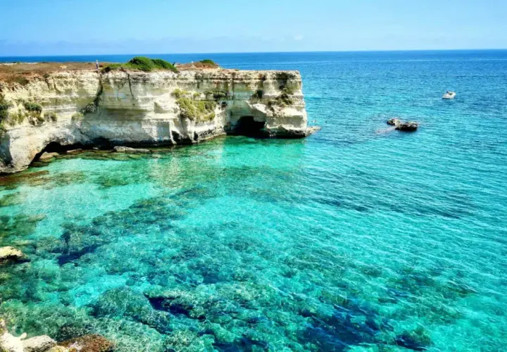 Turquoise water laps against white cliffs with caves, a boat sails in the distance.