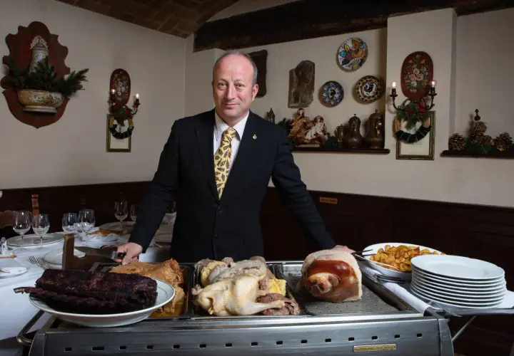 A man in a suit stands behind a cart with various meats, including ribs, a whole chicken, and a pig's head.