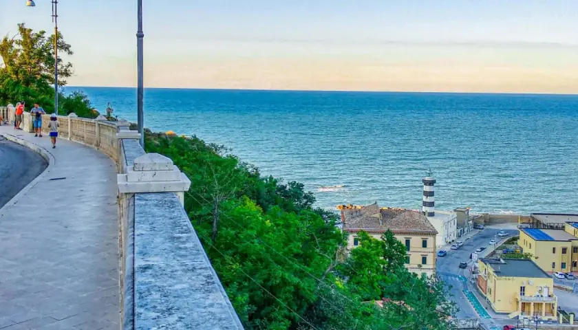 Coastal town with a lighthouse and buildings overlooking the blue sea, with a walkway in the foreground.