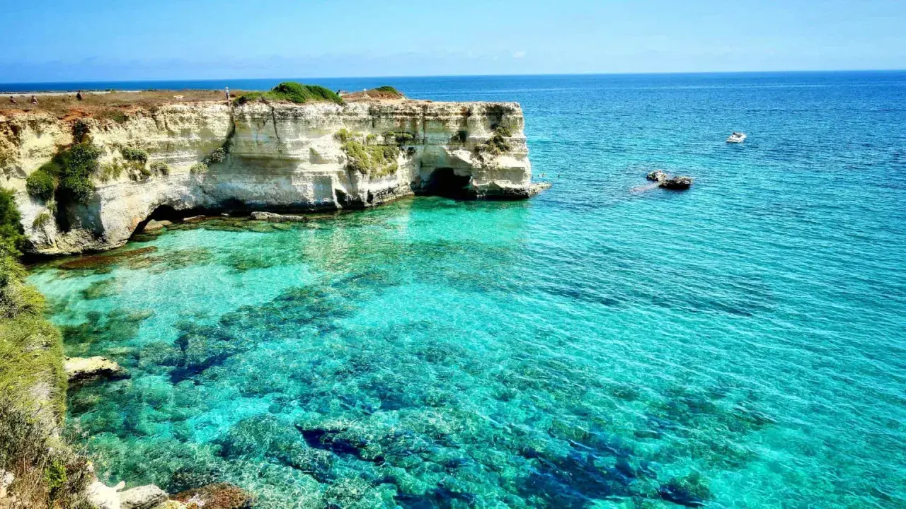 Turquoise water laps against white cliffs with caves, a boat sails in the distance.