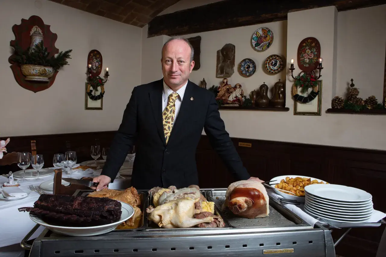 A man in a suit stands behind a cart with various meats, including ribs, a whole chicken, and a pig's head.