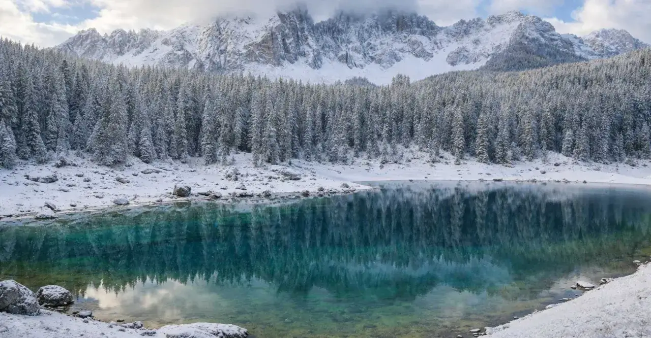 Lago alpino con riflessi di boschi innevati e montagne imbiancate. La neve in montagna crea uno scenario magico.