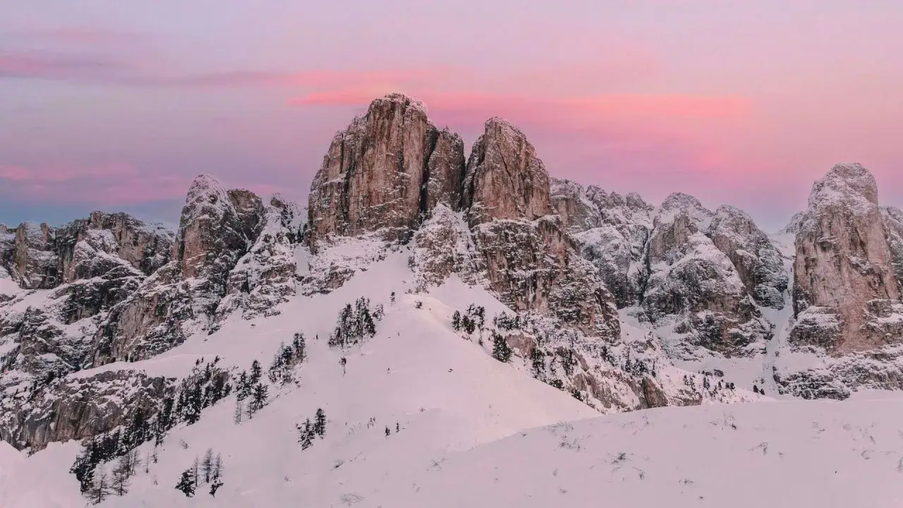 Cime rocciose innevate sotto un cielo rosa. La neve in montagna crea un paesaggio magico, con pini sparsi sui pendii.