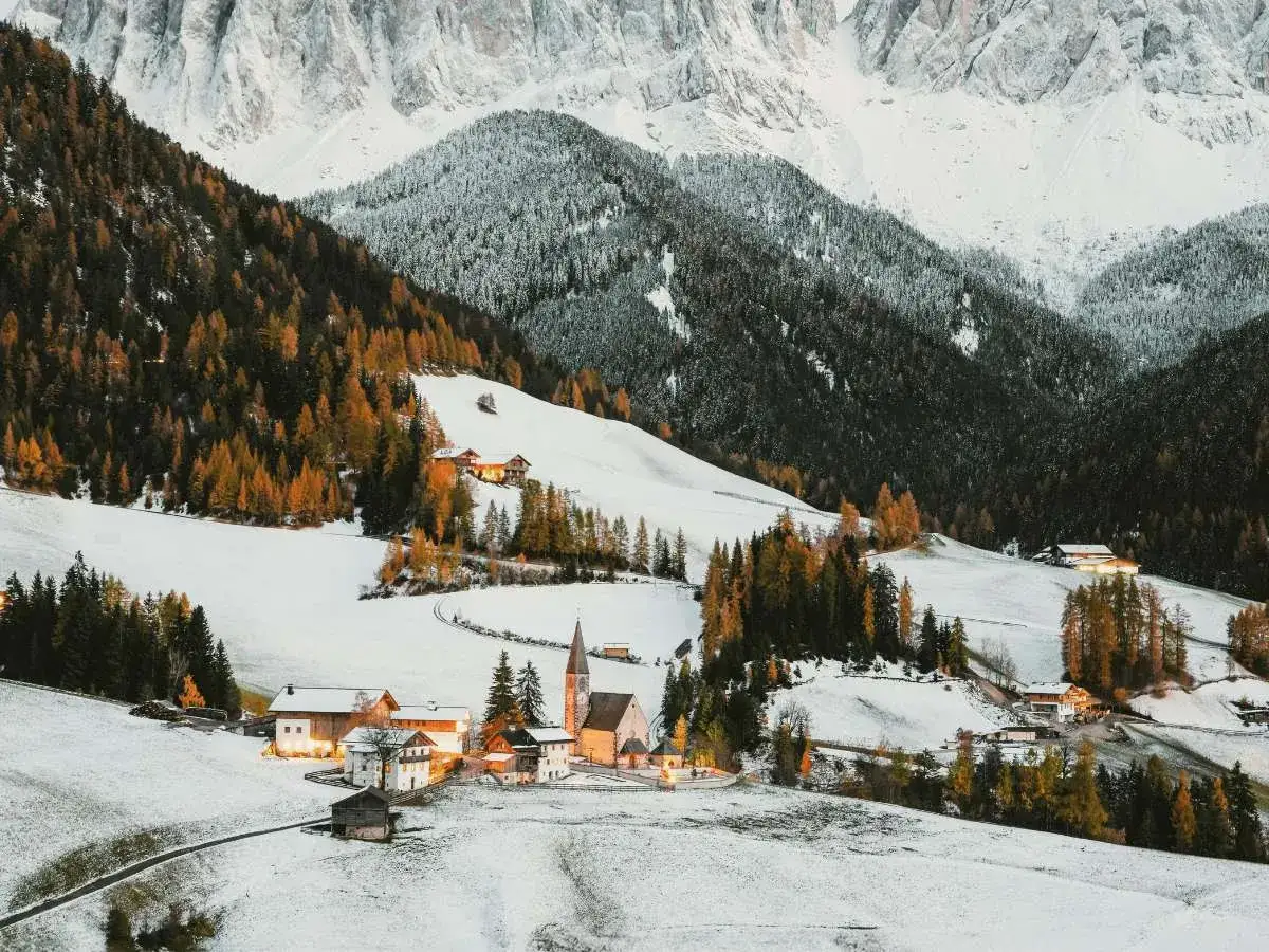 Un villaggio alpino imbiancato dalla neve, con case e una chiesa circondati da boschi autunnali e montagne innevate.